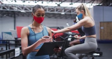 Jimnastik salonunda yüz maskesi takan karışık ırk kadın spor eğitmeni. Eğitmen, egzersiz bisikleti kullanarak yüz maskesi takan beyaz bir kadının zamanlamasını yapıyor. Coronavirus covid-19 salgını sırasında spor salonunda egzersiz yapıyor.