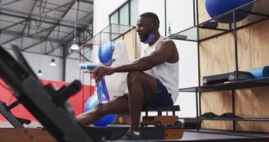 Spor salonunda spor yaparken maskesi takan Afrikalı Amerikalı bir adam. Mola vermek, su şişesini tutmak ve yüzünü havluyla silmek. Coronavirus covid 19 salgını sırasında spor salonunda hijyen.