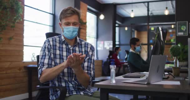 現代オフィスで手消毒を使用して顔マスクを身に着けている白人男性 コロナウイルスの職場での衛生と社会的距離は19のパンデミックを克服しました — ストック動画