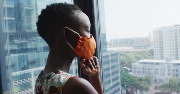 現代のオフィスで窓の外を見て顔のマスクを身に着けているアフリカ系アメリカ人女性の思考 コロナウイルスのパンデミック時の社会的距離隔離ロックダウン — ストック動画