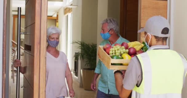 Entrega hombre entrega de comestibles a la pareja de ancianos caucásicos con máscaras faciales en casa — Vídeo de stock