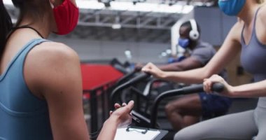 Spor salonunda kadın fitness antrenörü ve yüz maskesi takan bir müşteri. Koşu antrenörü beyaz bir kadının egzersiz bisikletini kullanmasını zamanladı. Coronavirus covid-19 salgını sırasında spor salonunda egzersiz yapıyor..