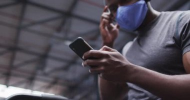 Spor salonunda akıllı telefon kullanarak yüz maskesi takan Afrikalı Amerikalı bir adam. Kulaklık takıyor, antrenman sırasında mola veriyor. Coronavirus covid 19 salgını sırasında spor salonunda egzersiz yapıyor..