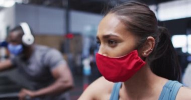 Yüz maskesi takan melez bir kadın spor salonunda egzersiz yapıyor. Çapraz antrenör makinesiyle çalışıyor. Coronavirus covid 19 salgını sırasında spor salonunda egzersiz yapıyor..