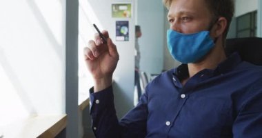 Yüz maskesi takan düşünceli beyaz adam modern ofisteki masasında kalem tutuyor. Coronavirus covid 19 salgını sırasında işyerinde hijyen ve sosyal mesafeler.