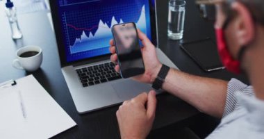 Modern ofisteki masasında otururken elinde akıllı telefon tutan beyaz bir adam. Koronavirüs salgını sırasında karantinaya alınma