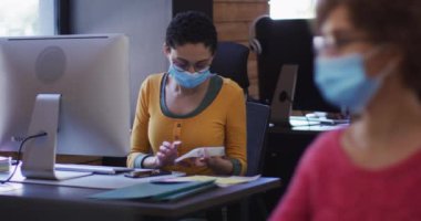 Yüz maskesi takan beyaz kadın belgeleri okuyor ve modern ofiste bilgisayar kullanıyor. Coronavirus covid 19 salgını sırasında işyerinde hijyen ve sosyal mesafeler.