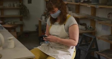 Çömlek atölyesinde akıllı telefon kullanan yüz maskesi ve önlük takan beyaz bir kadın. Coronavirus codemic 19 salgını sırasında çömlek atölyesinde hijyen ve sosyal mesafeler.