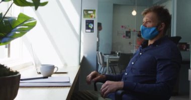 Yüz maskesi takan düşünceli beyaz bir adam dizüstü bilgisayar kullanıyor ve modern ofisin penceresinden dışarı bakıyor. Coronavirus covid 19 salgını sırasında işyerinde hijyen ve sosyal mesafeler.