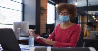 Modern ofiste dezenfektan sprey kullanarak akıllı telefonunu temizleyen yüz maskesi takan Afrikalı Amerikalı bir kadın. Coronavirus covid 19 salgını sırasında işyerinde hijyen ve sosyal mesafeler.