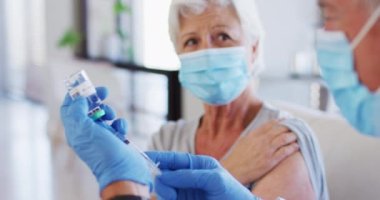 Beyaz erkek doktor şırıngayı ilaçla doldururken yaşlı beyaz kadın yüz maskesi takıyor.