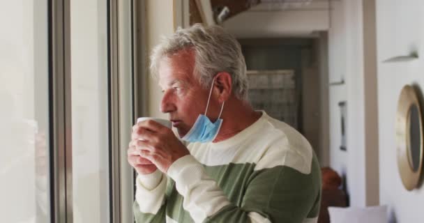 Starší běloch se sníženou obličejovou maskou popíjející kávu, zatímco se dívá z okna domů — Stock video