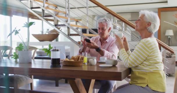 Starsza kaukaska para rozmawia ze sobą za pomocą smartfona jedząc razem śniadanie w domu — Wideo stockowe