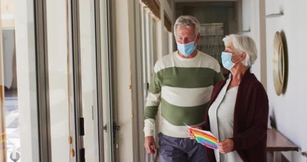 Starší kavkazský pár v obličejových maskách s duhovou malbou u okna doma — Stock video