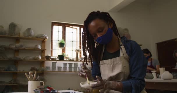 女陶工戴着面具和围裙 用刷子在陶器工作室的陶器上作画 大肠病毒大流行期间陶器工作室的卫生与社会疏离D — 图库视频影像