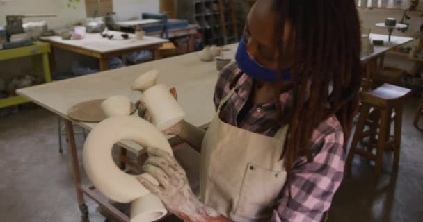女性アフリカ系アメリカ人の陶芸家が陶器工房でフェイスマスクとエプロンをつけています コロナウイルスの間の陶芸スタジオの衛生と社会的距離は19のパンデミックを克服しました — ストック動画