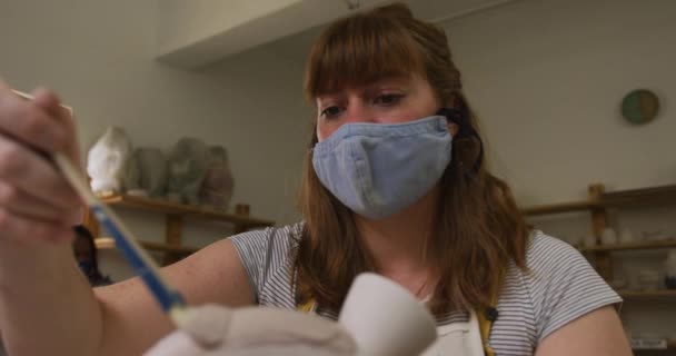 陶器の工房では 顔の面やエプロンをつけた女性陶芸家が釉薬を使って器に絵を描きます コロナウイルスの間の陶芸スタジオの衛生と社会的距離は19のパンデミックを克服しました — ストック動画