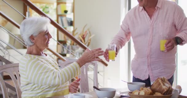 Senior caucásico pareja tostando vasos de jugo mientras desayunan juntos en casa — Vídeos de Stock