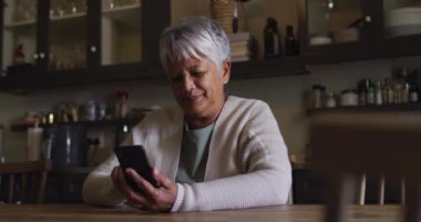 Gülümseyen son sınıf melez kadın mutfakta oturmuş akıllı telefon kullanıyor.