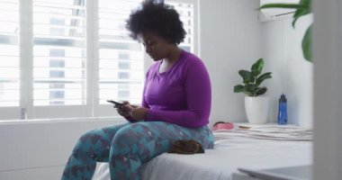 Afro-Amerikan kadın vlogger akıllı telefon kullanarak yatakta oturuyor.