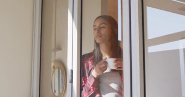 Karışık ırktan bir kadın. Pencere kenarında bir fincan kahve içiyor.