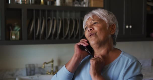 Feliz mujer de raza mixta senior hablando en el teléfono inteligente en la cocina — Vídeos de Stock