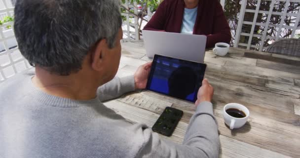Старшая смешанная раса пара сидит в саду с помощью ноутбука и планшета — стоковое видео