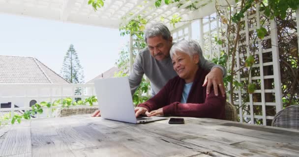 Feliz pareja de raza mixta senior sentado en el jardín utilizando el ordenador portátil — Vídeo de stock