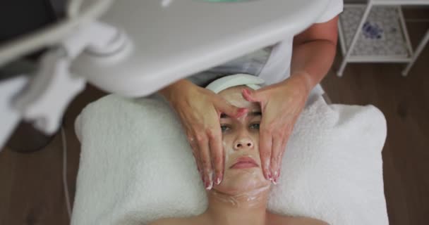 Biała kobieta leżąca z tyłu podczas gdy kosmetyczka robi jej maseczkę — Wideo stockowe