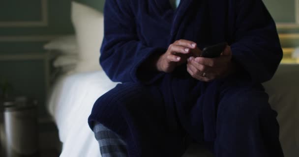 Hombre de raza mixta senior sentado en la cama usando teléfono inteligente, sección media — Vídeo de stock
