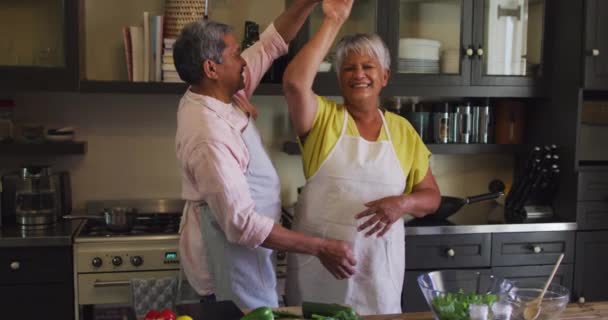 Щаслива старша змішана пара гонок у фартухах, що танцюють на кухні — стокове відео