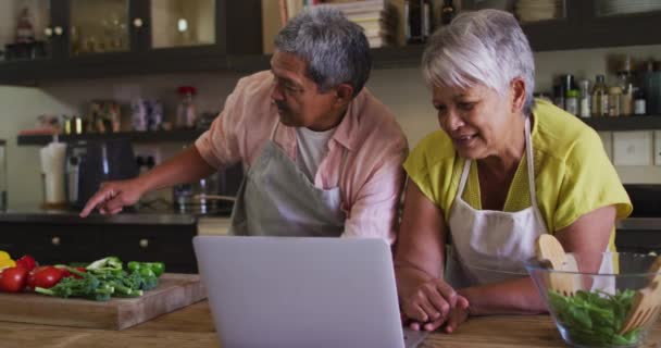 Pasangan ras campuran senior menggunakan laptop menyiapkan makanan di dapur — Stok Video