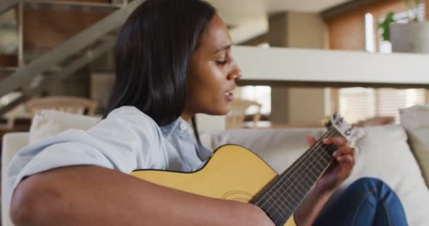 Μικτή γυναίκα φυλής στον καναπέ στο σπίτι παίζοντας κιθάρα και τραγουδώντας — Αρχείο Βίντεο