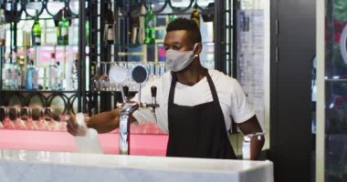 Yüz maskesi takan Afrikalı Amerikalı erkek barmen bar yüzeyini dezenfekte ediyor.