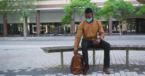 Hombre de negocios afroamericano con máscara facial sentado en el banco — Vídeos de Stock