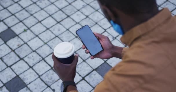 Αφροαμερικανός επιχειρηματίας φορώντας μάσκα προσώπου χρησιμοποιώντας smartphone κρατώντας καφέ — Αρχείο Βίντεο