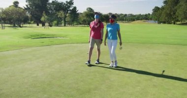 Yüz maskesi takmış golf oynayan iki beyaz kadın birlikte yürüyorlar.