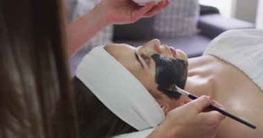 Güzellik uzmanı ona yüz maskesi takarken beyaz bir kadın sırt üstü uzanıyor.