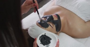 Güzellik uzmanı ona yüz maskesi takarken beyaz bir kadın sırt üstü uzanıyor.