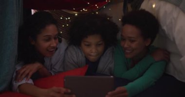 Yatak odası kampında yatan tablet kullanan melez lezbiyen çift ve kızı. Coronavirus covid 19 salgını sırasında evde tek başına vakit geçirmek..