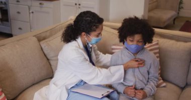 Karışık ırk doktoru maske takıp evde melez kızı muayene ediyor. Bir kanepede steteskop kullanarak oturmak. Coronavirus covid 19 salgını sırasında evdeki kişisel izolasyon tıbbi iletişimi.