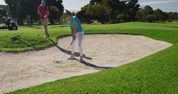 Två kaukasiska kvinnor spelar golf i ansiktsmasker en tar skott från bunkern — Stockvideo