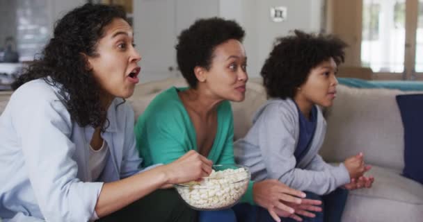 ポップコーンを食べるテレビを見ている混合レースレズビアンのカップルと娘 興奮して応援に飛び込む 家庭での自己分離の質家族の時間コロナウイルスCovid 19パンデミックの間 — ストック動画