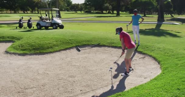Two caucasian women playing golf wearing face masks one taking shot from bunker — Stock Video