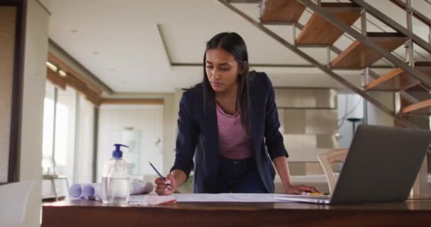 Mixed race woman by desk at home using laptop looking through blueprints — Stock Video