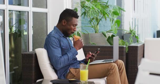 Hombre de negocios afroamericano usando computadora portátil, teléfono inteligente comer croissant en la cafetería — Vídeo de stock