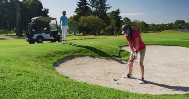 Två kaukasiska kvinnor spelar golf i ansiktsmasker en tar skott från bunkern — Stockvideo