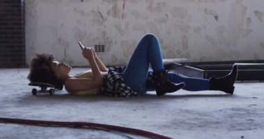 Karışık ırk kadını boş bir binada kaykayın üstünde uzanmış, akıllı telefon kullanarak gülüyor.