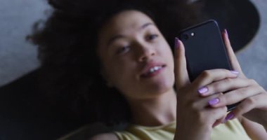 Karışık ırk kadını boş bir binada kaykayın üstünde uzanmış, akıllı telefon kullanarak gülüyor.