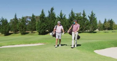 Golf sahasında golf çantalarıyla yürüyen yüz maskesi takan iki Kafkasyalı son sınıf öğrencisi. Coronavirus covid 19 salgını sırasında emeklilik sporları ve aktif yaşlı yaşam tarzı.